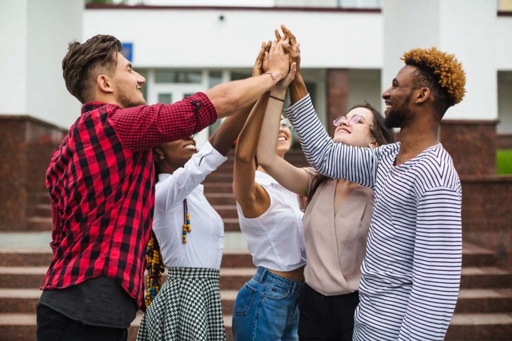 Gírias mais comuns da língua inglesa Cinco amigos jovens, sendo um rapaz branco de cabelos escuros e blusa xadrez vermelha e preta, uma menina negra de blusa branca e saia xadrez preta e branca, uma menina de óculos com blusa branca e calça jeans azul, uma menina de blusa bege e calça preta e um rapaz negro com blusa listrada de azul e branco - estão tocando as mãos.