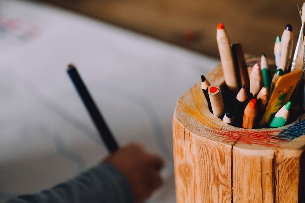 Criança desenhando ao fundo de um copo com vários lápis de cor, representando a cultura maker na prática.
