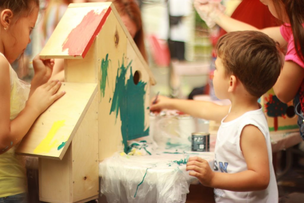 Dicas para estimular a criatividade infantil no cotidiano Duas crianças se reúnem para pintar uma casinha de madeira, na imagem, em uma atividade em família para exercitar a criatividade infantil.