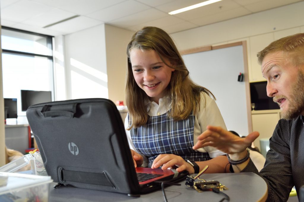 Garota adolescente recebendo uma aula nos moldes da disciplina positiva, usando um computador enquanto o professor dá explicações.