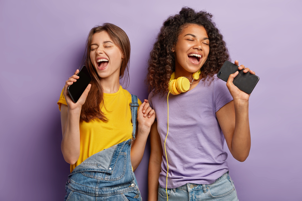 Duas garotas segurando seus celulares, que simulam microfones. As duas estão de olhos fechados cantando músicas em inglês.