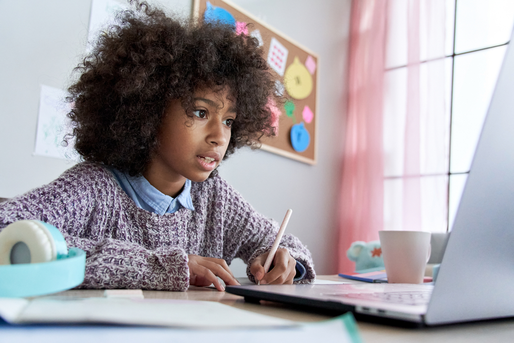 Criança anotando em caderno o que vê em seu laptop: está estudando sobre os costumes da língua inglesa e suas expressões idiomáticas.